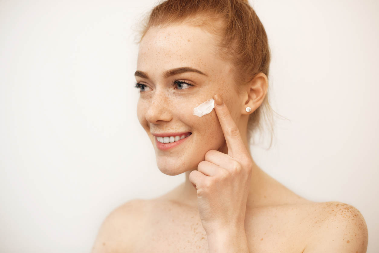 La belle fille caucasienne avec les cheveux roux et les taches de rousseur sourit tout en utilisant une crème sur son visage