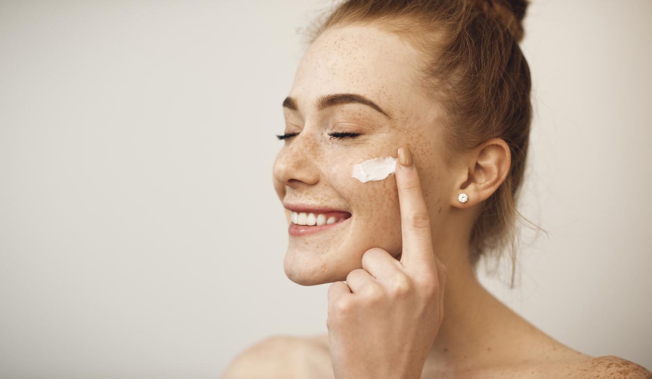 jeune femme avec les cheveux roux et les taches de rousseur appliquant la crème blanche sur son visage riant